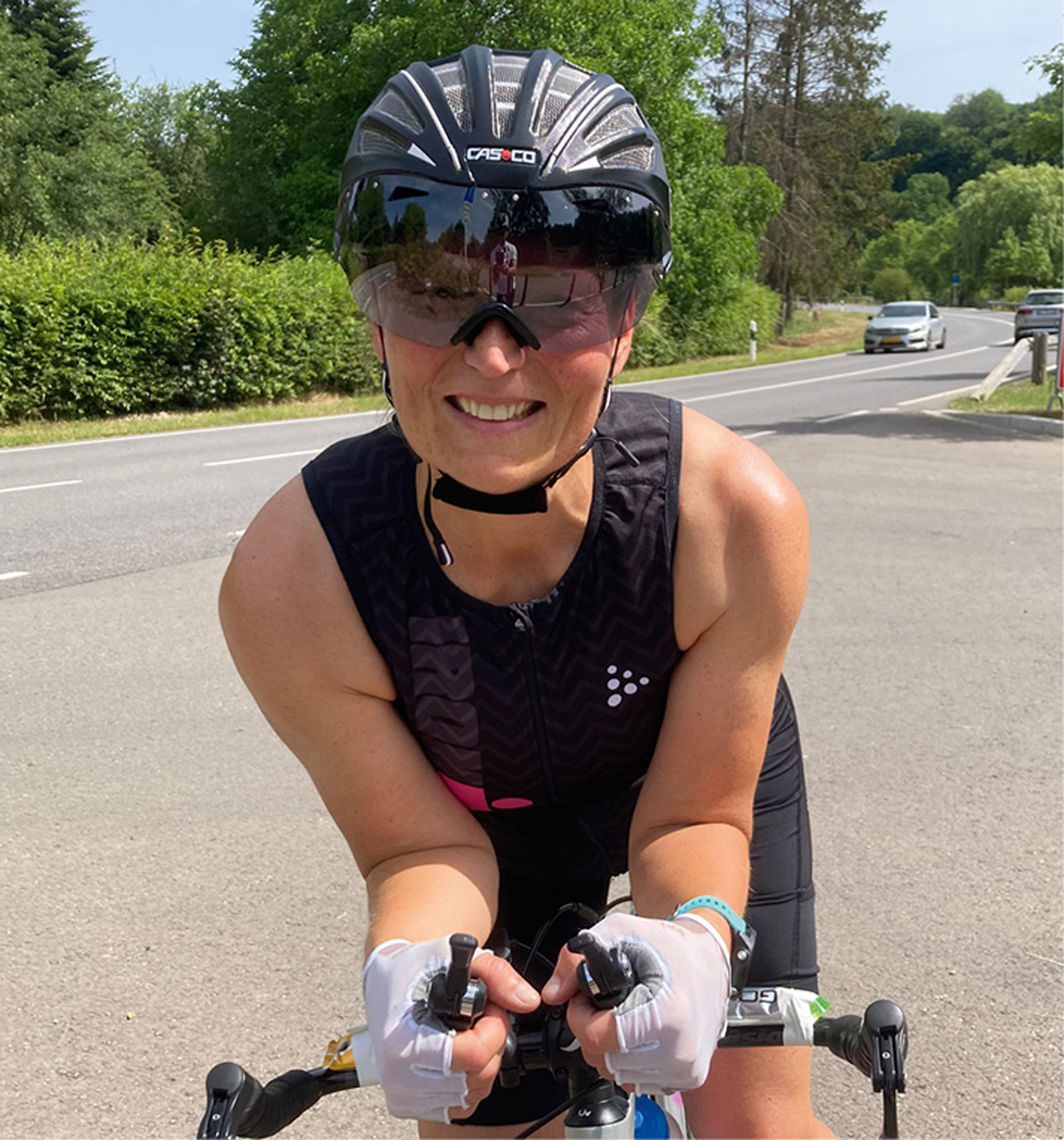 Triathletin in Triathlon-Ausrüstung bei Sommertraining auf Landstraße Triathletin mit Casco-Helm und Sonnenbrille auf Rennrad, auf einer sonnigen Straße mit grüner Landschaft